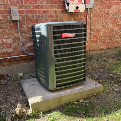 A Goodman air conditioning unit sits on a concrete slab beside a brick wall, with utility panels visible in the background.