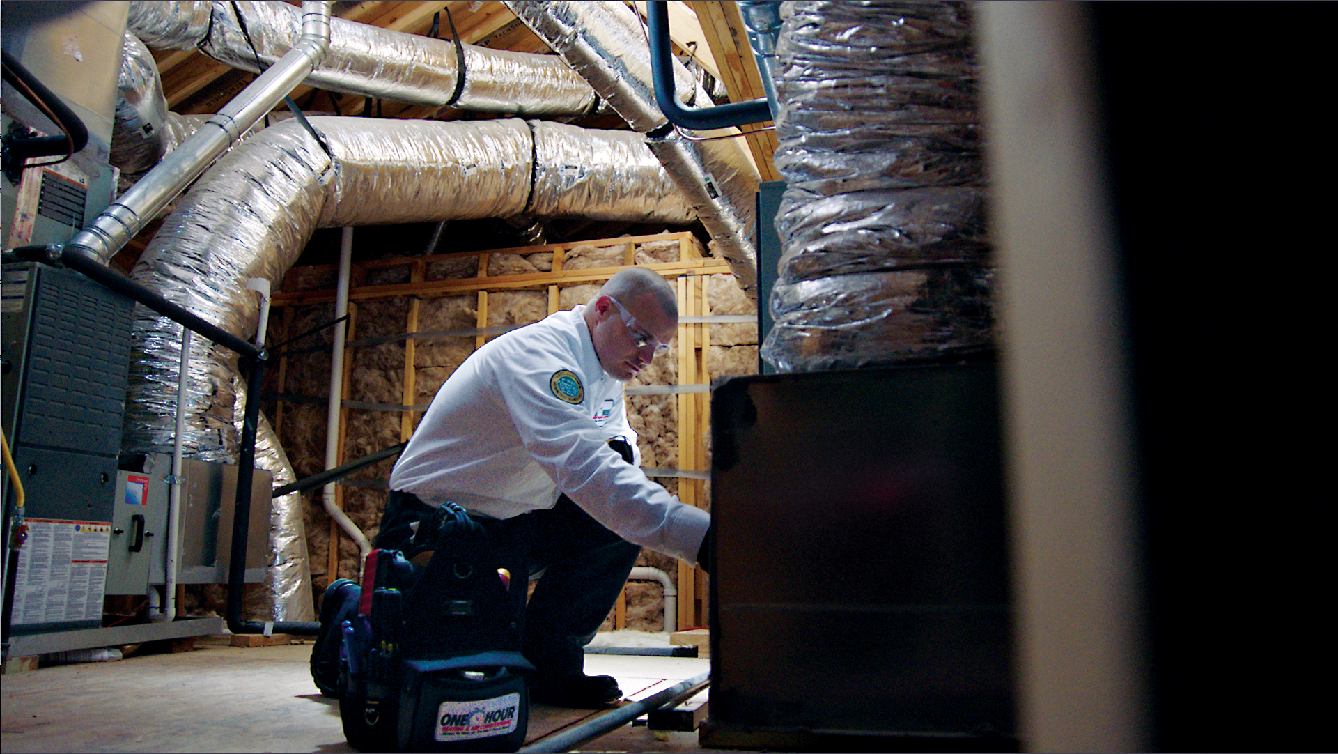 HVAC technician performing furnace maintenance inside an attic with ductwork and insulation visible, photographed for One Hour Heating & Air Celina.